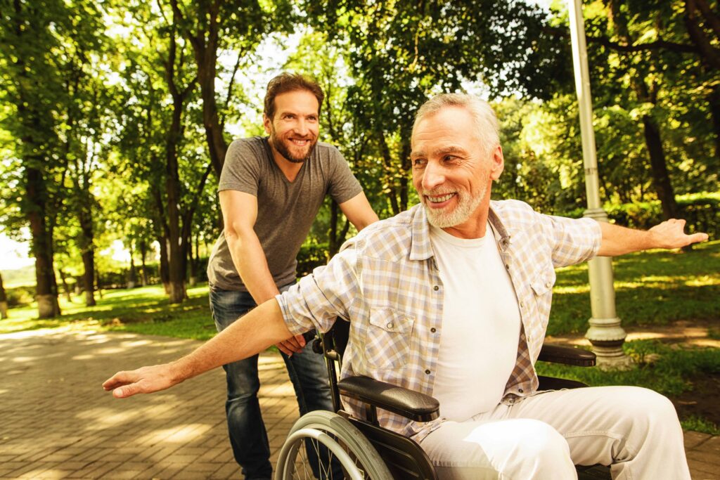 The old man on a wheelchair is walking in the park with his adult son